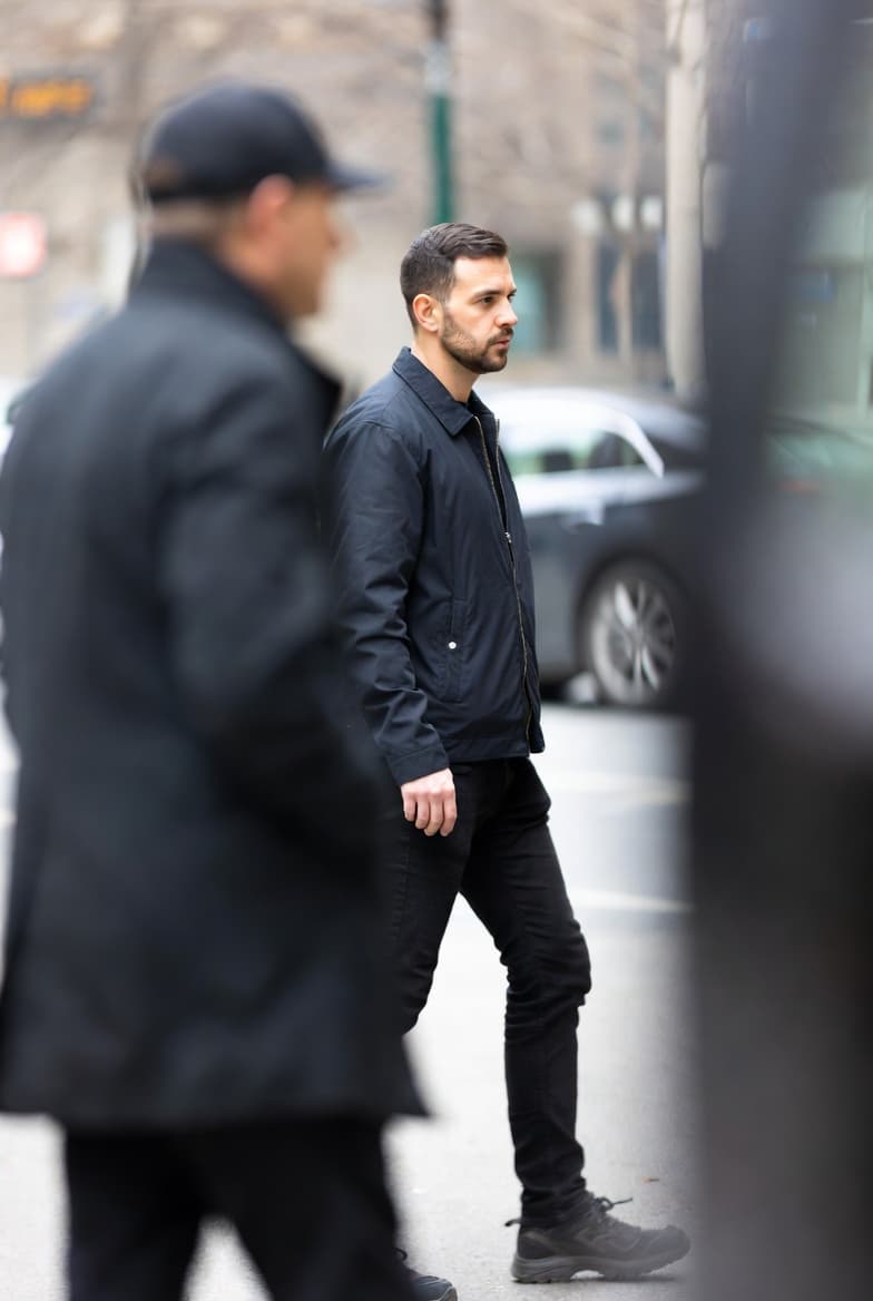Bearded man in a navy jacket and black pants walking on a city street.
