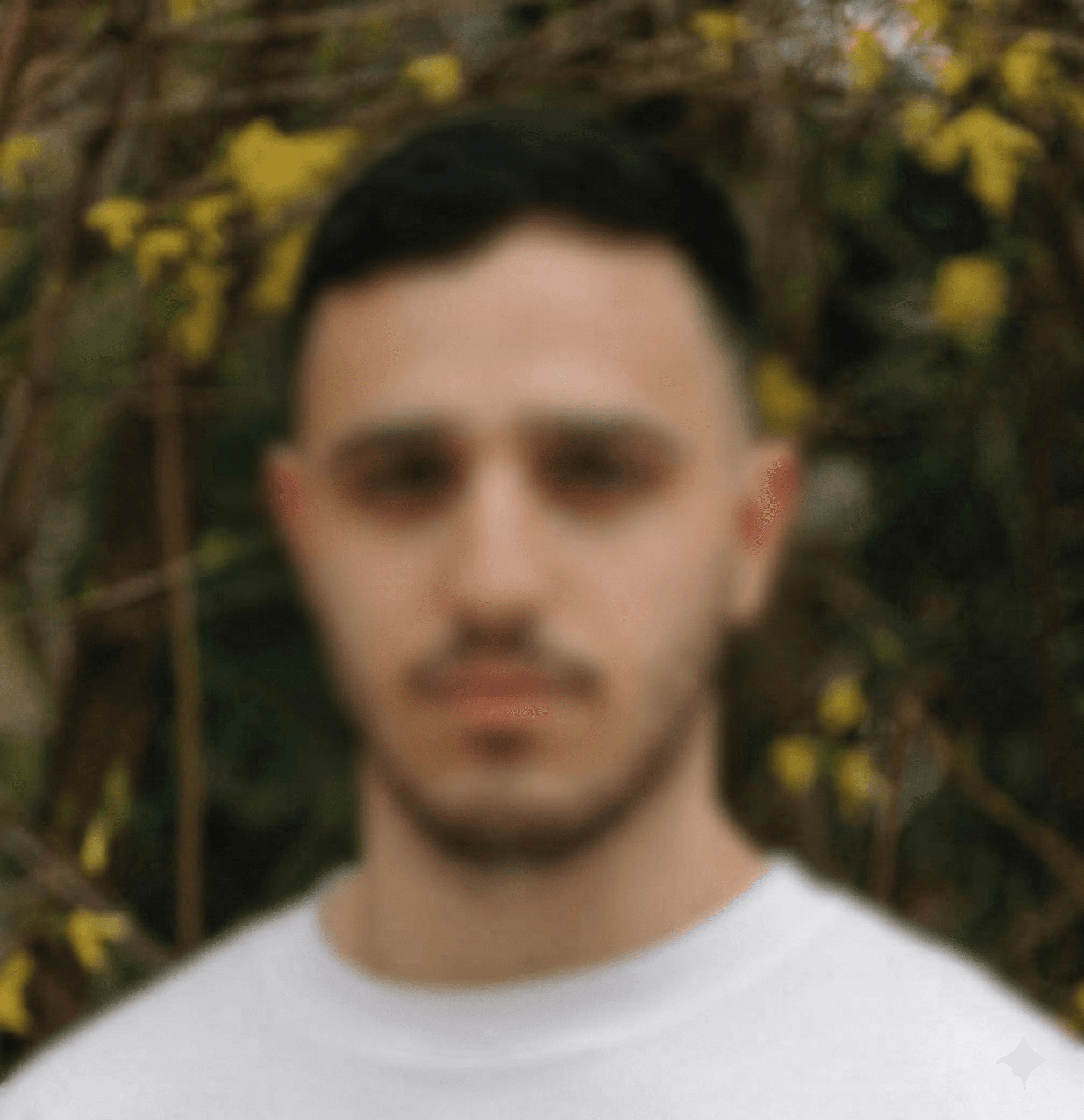 Blurry portrait of a young man with dark hair and beard wearing a white shirt.