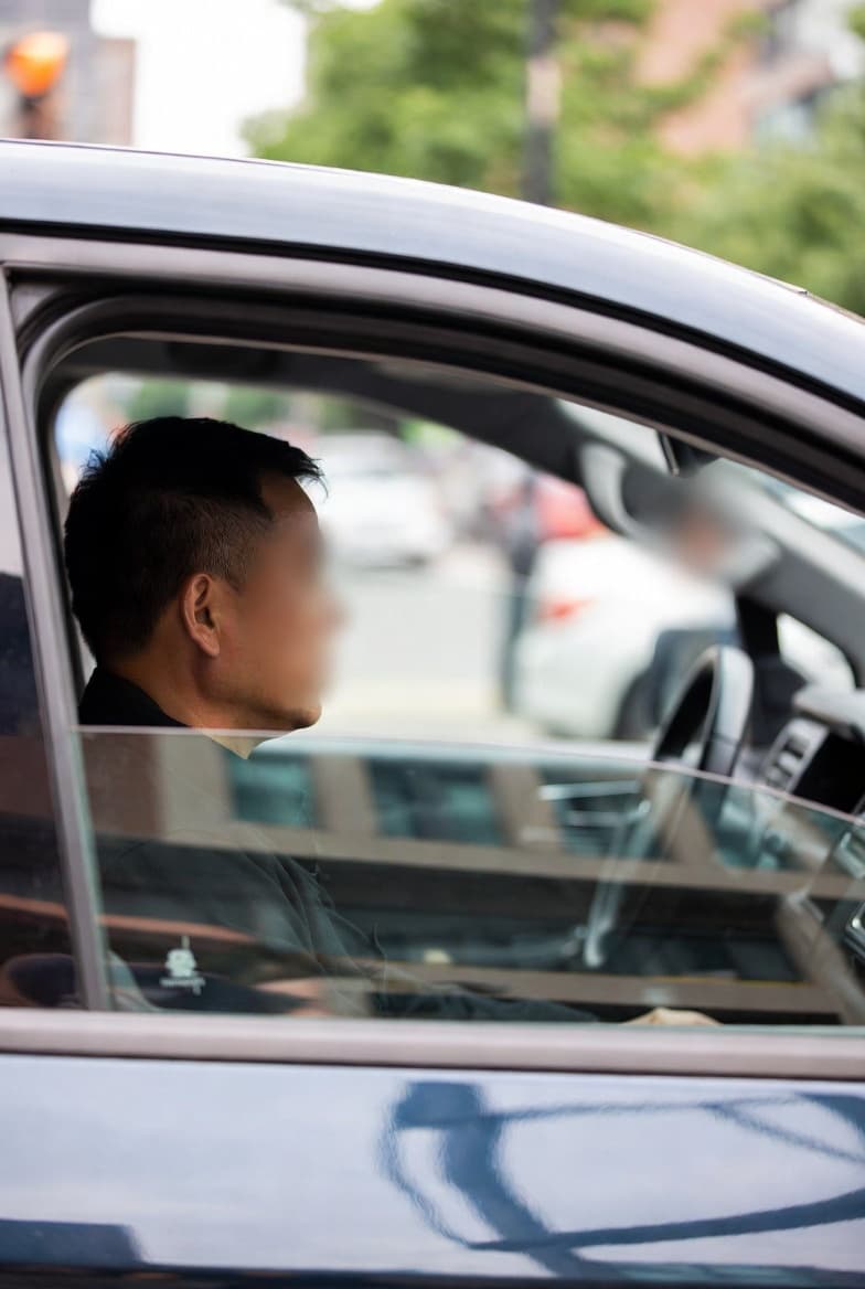 Side profile of a man with short dark hair sitting in a car, face blurred.